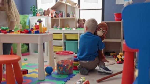 Woman smiles while boy holds a plush monkey and plays with building blocks at a playroom table in building; playful bonding.