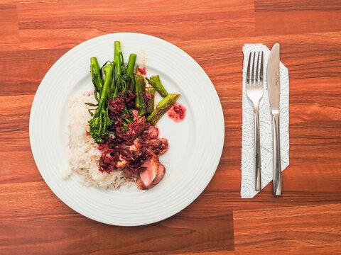 White plate holds roasted duck breast, rice, and green vegetables. Dish rests on wooden table. Fork and knife sit beside plate. Food looks freshly prepared and ready to eat