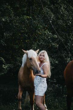 A blonde woman walks with horses in the garden in summer