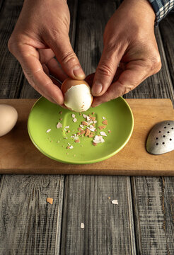 Hands are seen breaking an egg over a green bowl. Eggshells and bits of shell are visible on the wooden table next to the bowl. Other eggs are on the table