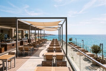 Obraz premium Spacious waterfront dining terrace with rows of wooden tables, sun shade sails, glass railing, and calm blue sea beyond rocky coastline, showing bright daylight and relaxed hospitality setting
