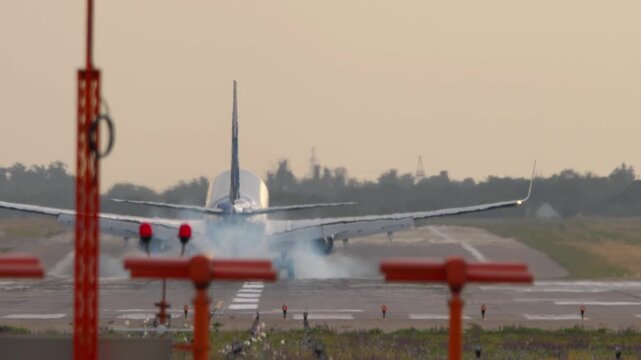 Passenger jet touching down on airport runway with tire smoke at sunset, featuring heat haze, red approach lights in foreground, telephoto rear view
