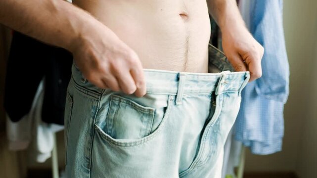 Man shows weight loss and body transformation by holding out loose, oversized jeans from his waist. A perfect concept of healthy habits and diet success. High quality 4k footage