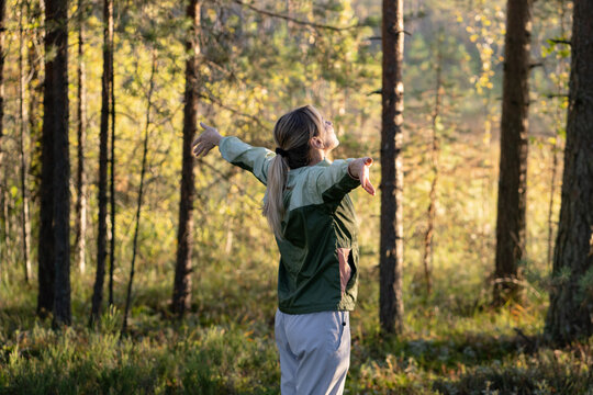 Cheerful woman embracing nature on walk in forest, standing with spread arms inhaling fresh air, positive calm female taking deep breath of air with raised hands, health therapy and mindfulness.