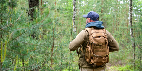 Adult man with backpack stands on forest trail with back to camera and looks ahead among tall pines. Hiking, travel and recreation concept © Dmitrii Masquerade