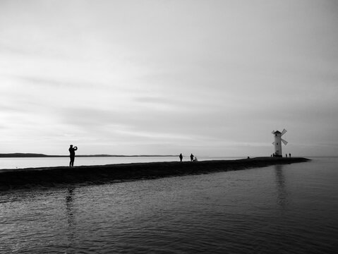 A black and white image shows a long pier extending into the water with a lighthouse at the end. Several people are visible on the pier, some walking and one taking a photo. Stawa Mlyny, Poland. 