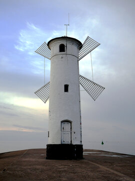 A white, cylindrical windmill with four prominent sails stands on a stone pier against a cloudy sky. The windmill has a dark base and a single door near the bottom. Stawa Mlyny, Poland. 