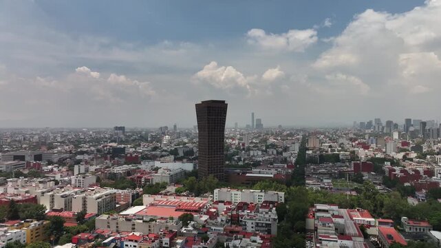 Aerial panoramic view of the Mexico City skyline looking south with a prominent skyscraper, drone flying backward over the sprawling cityscape under a cloudy blue sky
