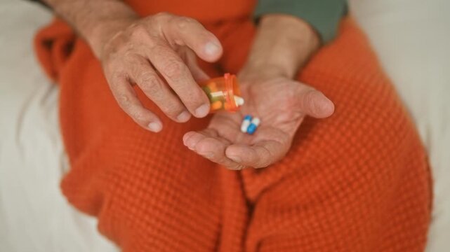 Man offering blue and white capsules on bed in building, open palm visible and pillbottle nearby; vulnerability solitude.