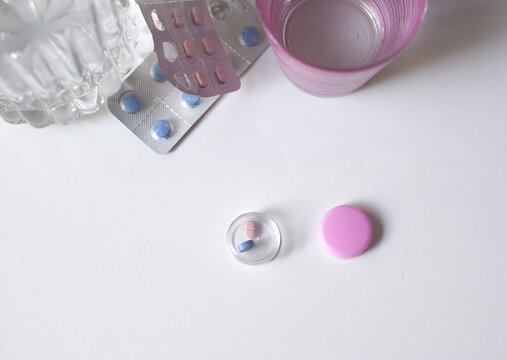 Pastillero o contenedor con una pildora azul y una roda sobre una mesa blanca, junto a un blister de pastillas azules y rosas, vaso y jarra