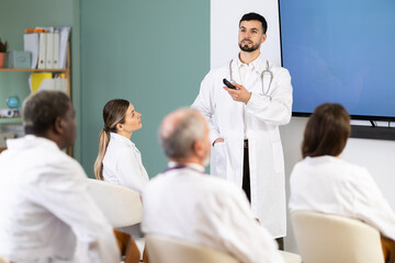 Fototapeta premium Young male doctor in medical gown with remote control gives presentation to colleagues doctors in conference room
