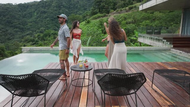 Group of young people dancing by the pool at a small party on wooden deck with cocktails. Friends enjoy music near infinity pool overlooking lush green mountains at luxury villa in slow motion.