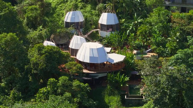 Aerial view of a luxury villa in Costa Rica featuring unique pod-shaped structures with white roofs nestled among dense tropical rainforest. Wooden walkways connect elevated glamping units with pool.