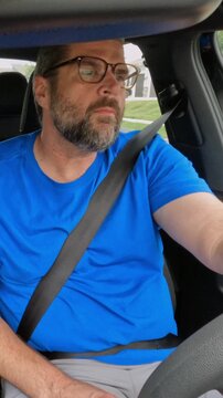 Middle aged man wearing blue shirt and glasses driving compact car, looking out the window and stretching his sore neck from long distance driving
