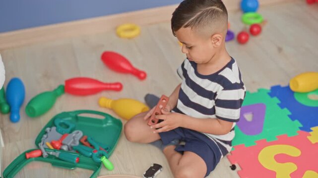 Little boy sitting on playroom floor holding toy toolkit pieces with striped shirt visible in studio; quiet concentration.