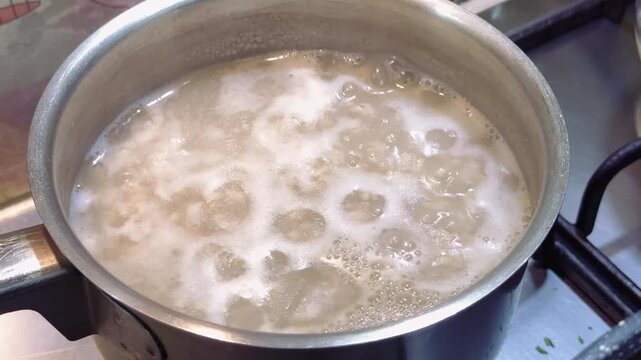 Pearl barley porridge is cooking in a stainless steel pan on a gas stove. Bubbles form as the grains absorb water, creating a creamy texture