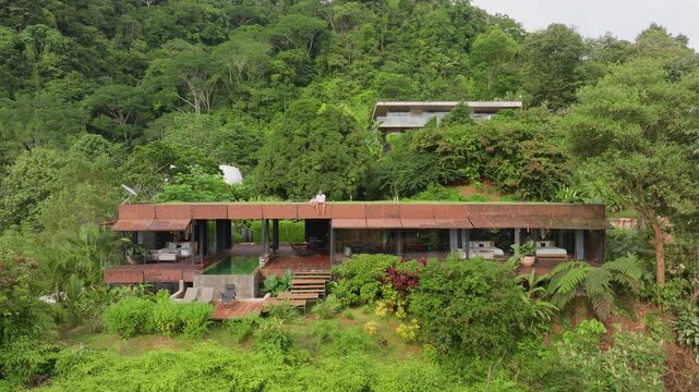 Aerial drone footage captures modern architectural villa with weathered steel roof and glass walls surrounded by dense green tropical jungle on hillside with couple sitting on rooftop terrace.
