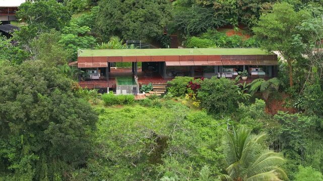 Aerial view of a stunning modern luxury villa with green living roof and weathered steel exterior nestled into lush tropical rainforest hillside with pool deck and open-air living spaces visible.