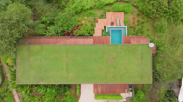 Aerial drone footage captures a luxurious tropical villa in Costa Rica featuring a turquoise swimming pool, wooden deck terraces, manicured green lawn, and lush jungle surroundings on an overcast day.