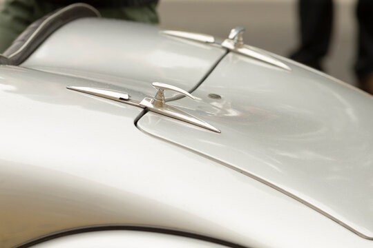 Shallow depth-of-field close view of a polished silver vintage car hood featuring twin streamlined chrome hood ornaments and smooth curved body lines. Soft