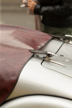 Detailed close-up of a vintage car hood showing a worn reddish-brown leather bonnet cover secured by chrome hood fasteners and straps. The pale silver painted