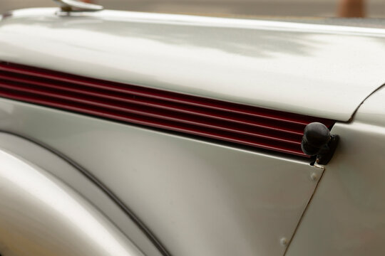 Detailed close-up of a vintage car hood showing cream-colored painted metal, curved fender edge, and horizontal red louver vents. Visible rivets and a black