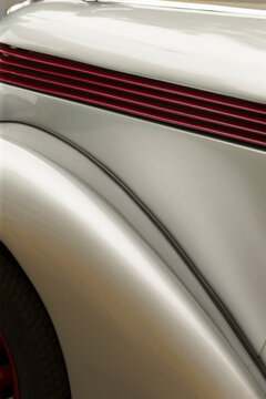 Tight detail view of a light-colored vintage car showing the curved front fender, hood area, horizontal red grille slats and subtle chrome trim. Soft studio