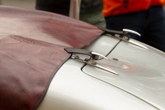 Close-up view of a leather tonneau cover secured over the front of a classic silver car by polished chrome hood latches and clamps. The image shows textured