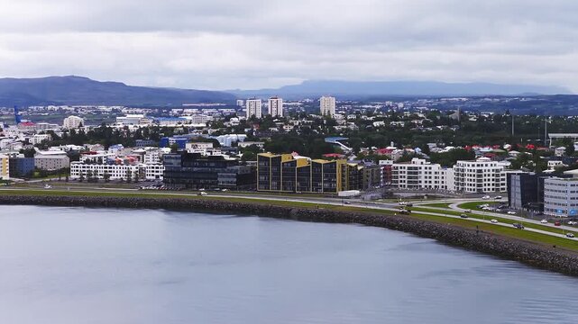 Aerial sweep along Reykjavik waterfront shows curved seawall, calm bay, yellow accented apartments, coastal ring road traffic, Hallgrimskirkja and Esjan under overcast skies.
