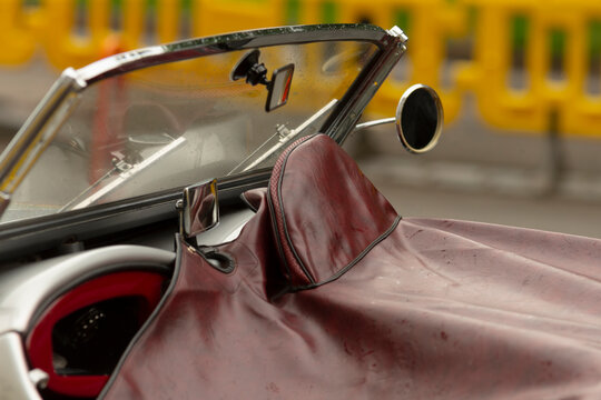 Tight view of a vintage convertible's cockpit featuring a burgundy leather tonneau cover stretched over the passenger area, a small round side mirror, and a
