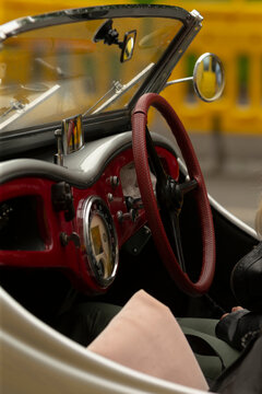 Close-up view of a vintage convertible car interior featuring a textured red steering wheel, classic dashboard with round chrome-trimmed gauges, partial