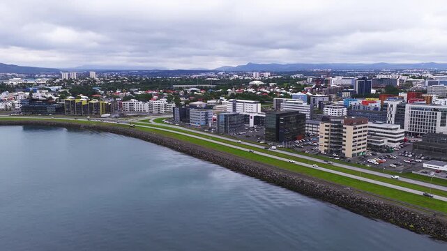 Aerial view of Reykjavik waterfront with curved seawall and ring road, modern glass blocks, mid rise apartments, colorful houses, and Esjan on the horizon.
