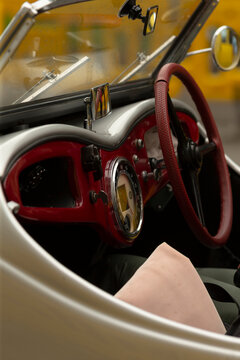 Close-up view of a vintage convertible car interior showing a glossy red dashboard, chrome-trimmed round gauge, textured leather-wrapped steering wheel in