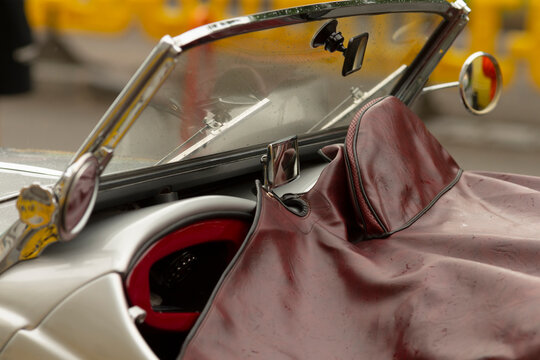 Close-up view of a classic convertible's cockpit featuring a chrome-framed windshield, leather tonneau cover draped over the passenger area, padded leather