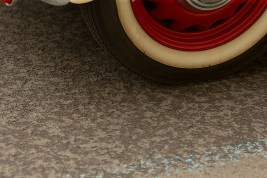 Low-angle view showing a vintage car wheel with a whitewall tire and glossy red steel rim resting on rough textured asphalt. Soft directional lighting
