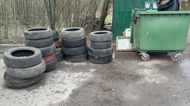 Green dumpster and stacked tires on gritty roadside during spring day, worn rubber wheels piled beside municipal container