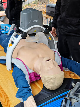 Lucas 3 automated chest compression device on CPR mannequin, vertical closeup at outdoor emergency medical demonstration, mechanical resuscitation robot for first aid and paramedic training