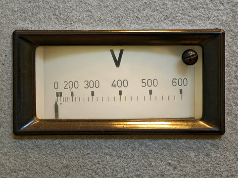 Old analog voltmeter gauge with needle at zero, front view closeup of vintage voltage meter scale from 0 to 600 volts in brown frame on rough gray panel, electrical measurement