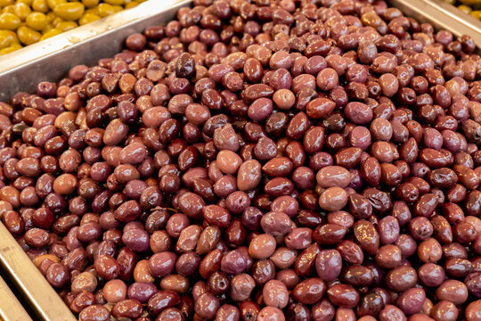 Heap of glossy purple olives in stainless steel market tray, closeup of brined kalamata style deli food with green olives behind, Mediterranean antipasti, organic grocery and snack concept, no people