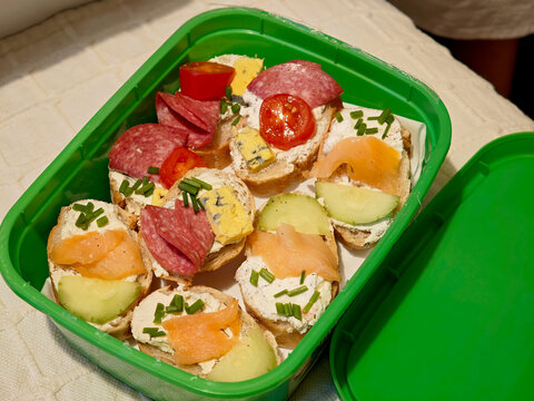 Homemade mini open sandwiches in green plastic container, top angle closeup of party canapes with smoked salmon, salami, cream cheese, melon cherry tomato and chives for catering picnic or movie night