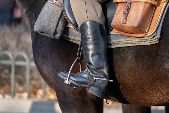 Anonymous rider boot in metal stirrup on dark horse, closeup of saddle, leather pouch and equestrian gear, cavalry reenactment, mounted patrol, horseback riding tradition and horsemanship scene detail