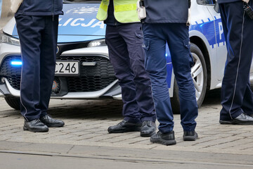 Anonymous Polish police officers standing beside patrol car with blue emergency lights, cropped uniformed legs at street gathering, law enforcement patrol, public safety, crime scene and news concept © Tomasz