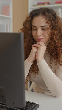 Woman clasping hands under chin at computer monitor in building while studying the screen; calm concentration.