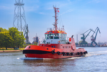Fototapeta premium Red harbor tugboat navigating a waterway near the Gdansk Shipyard with industrial cranes and port infrastructure in Gdansk, Poland.