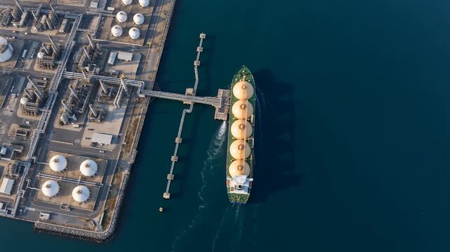 Aerial top view of an LNG carrier moored at a natural gas terminal loading jetty, overhead drone shot of a gas tanker docked at a liquefied natural gas plant with pipelines