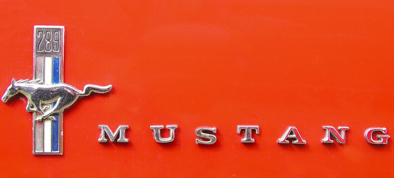  Close-up of a chrome Ford Mustang running horse emblem and the word Mustang mounted on a bright red vehicle highlighting automotive design, vintage styling and motion inspired branding.