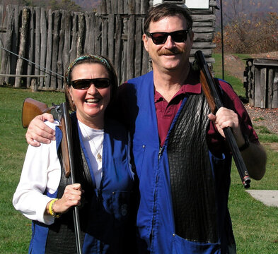 Smiling couple wearing shooting vests pose with shotguns at a country shooting range, enjoying outdoor recreational clay target shooting and camaraderie on a sunny autumn day.