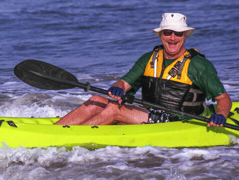 A smiling senior man paddles a bright yellow kayak through surf near shore wearing a life jacket, sun hat and sunglasses, enjoying outdoor adventure, summer fun and active coastal recreation.