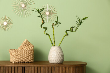 Fototapeta premium Vase with bamboo stems and wicker baskets on wooden cabinet near green wall