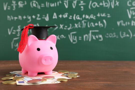 Piggy bank with graduation hat and money on table near green blackboard. Student loan concept